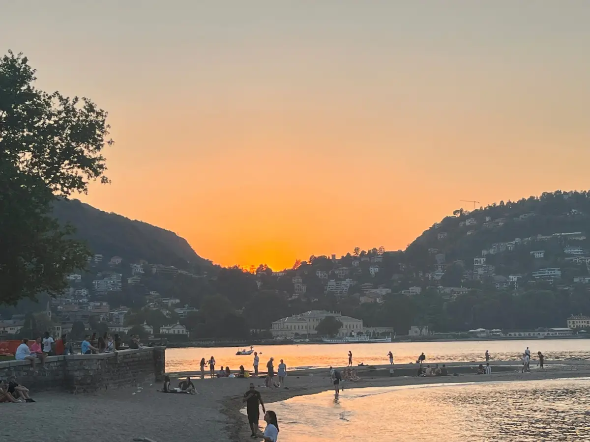 Ηλιοβασίλεμα στη Λίμνη Κόμο με κόσμο να περπατά και να χαλαρώνει στην παραλία, απολαμβάνοντας τη θέα και την ατμόσφαιρα του καλοκαιριού.
