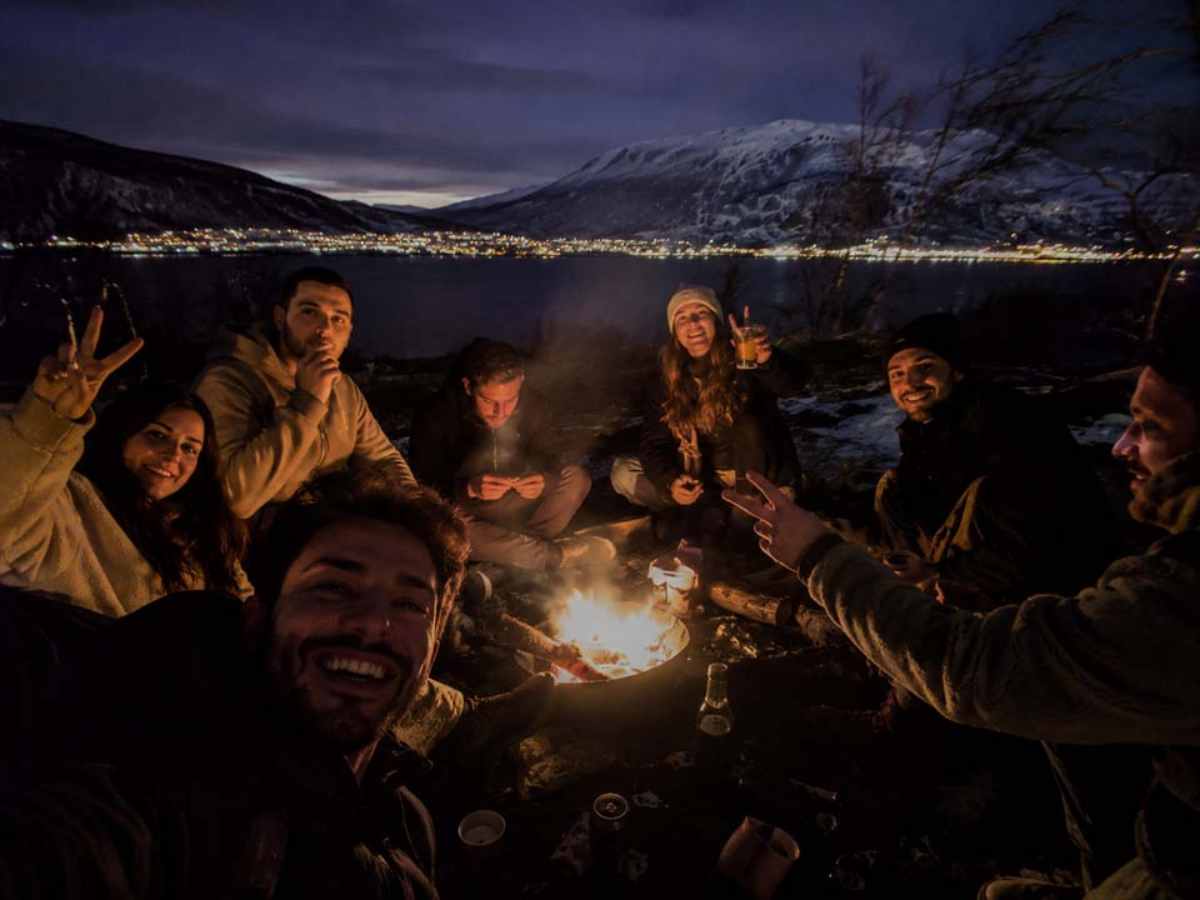 Παρέα ταξιδιωτών γύρω από φωτιά δίπλα σε λίμνη σε βραδινό χειμερινό roadtrip